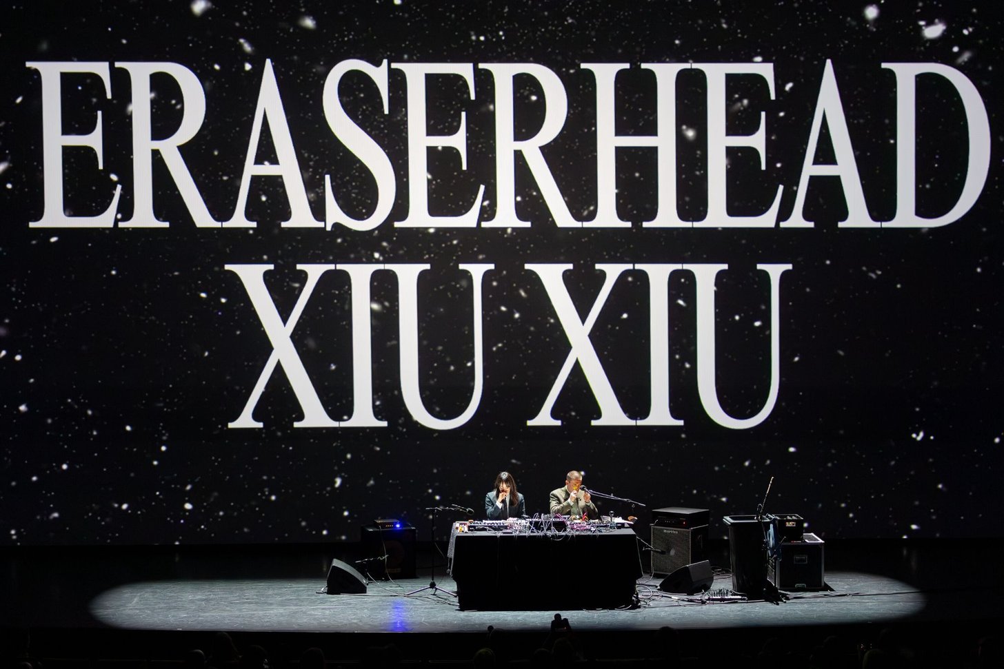 ERASERHEAD XIU XIU projectado sobre um campo de estrelas, dois intérpretes à mesa.