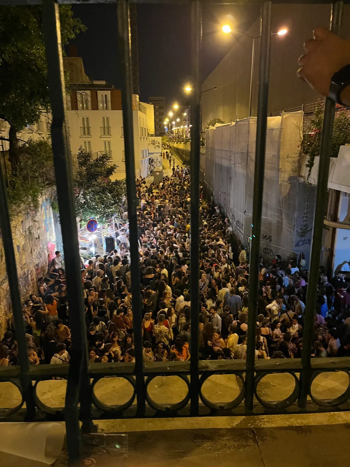 Multidão nocturna vista através de um gradeamento, sob os candeeiros da rua.