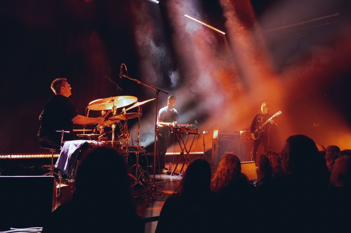 Uma banda em palco entre fumo laranja: bateria, mesa de efeitos e baixo.