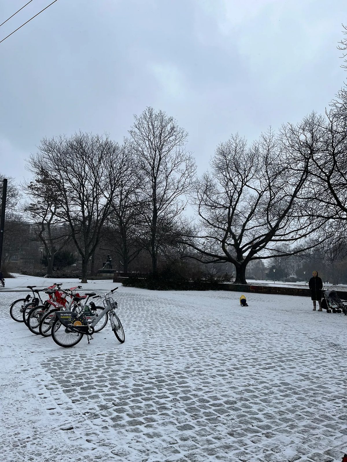 Praça coberta de neve, com bicicletas e árvores despidas.